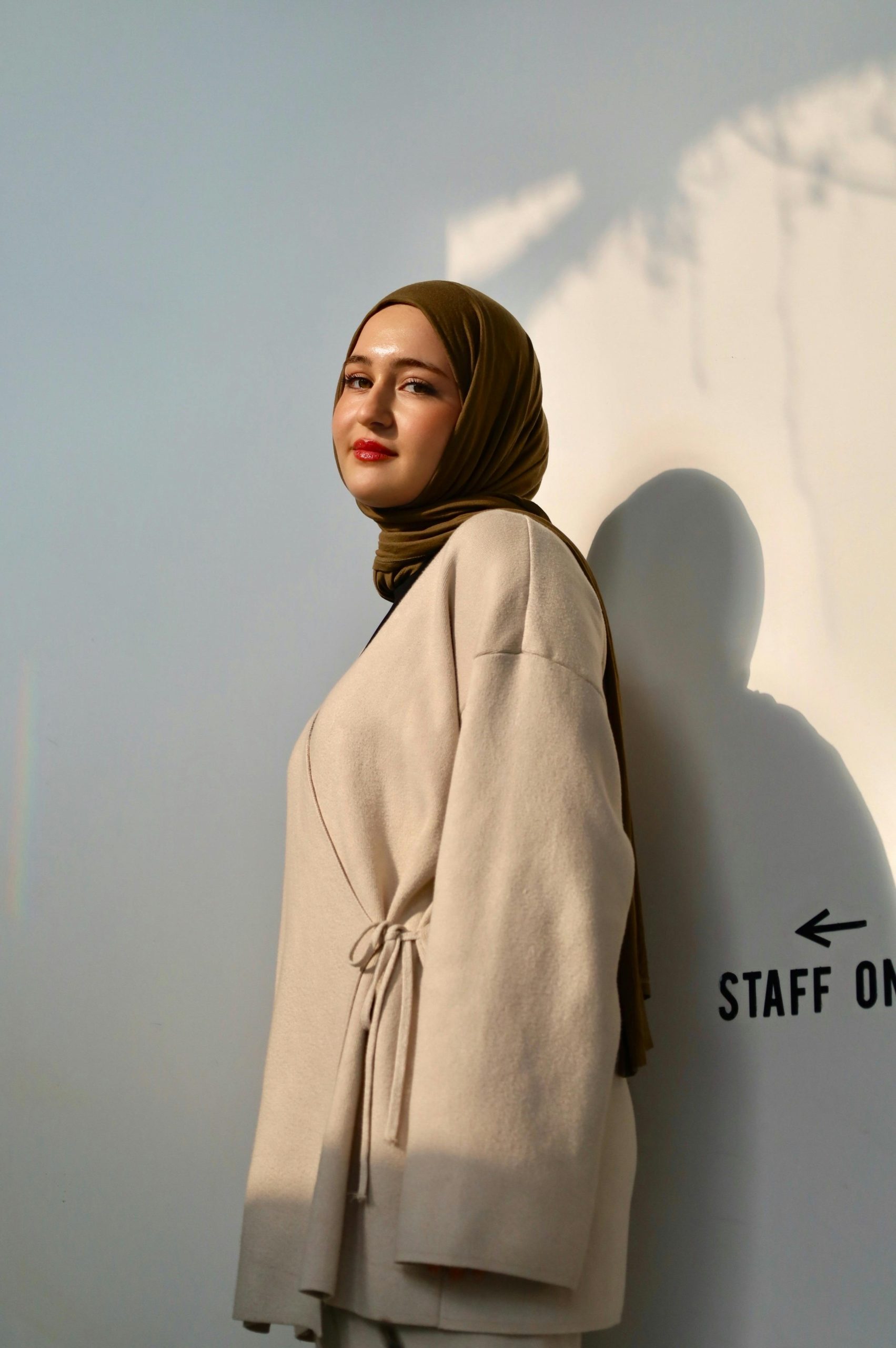 Profile portrait of a fashionable young woman in a hijab against a sunlit wall indoors.